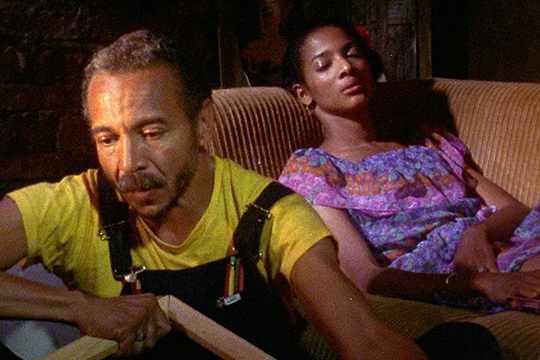 Light skinned man in yellow shirt sitting on ground. To his right is a dark skinned woman in a purple top laying on a couch and staring at camera
