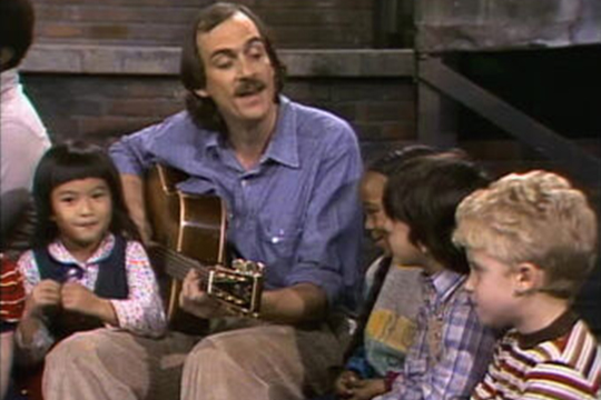 Man with long hair and mustache playing a guitar looking down at a blond kid with a striped shirt and a kid with brown hair to his left