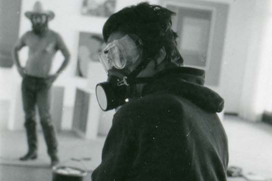 Lynda Benglis stands in studio with gas mask on while man stand akimbo with cowboy hat on
