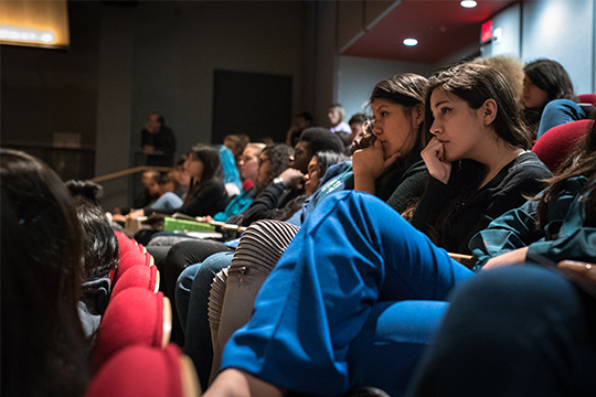Audience members listen intently to a lecture while seated in an auditorium.