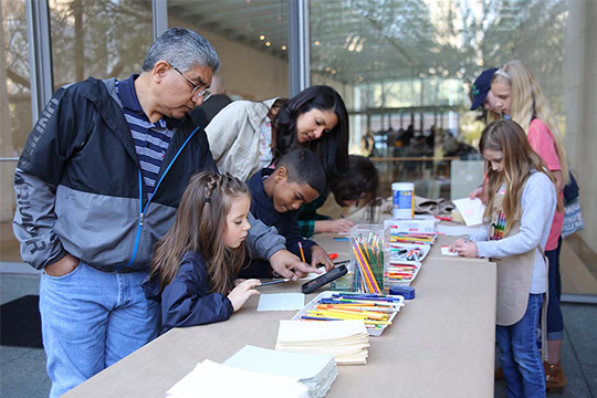 Diverse families work at an outdoor art station with colored pencils and paper.