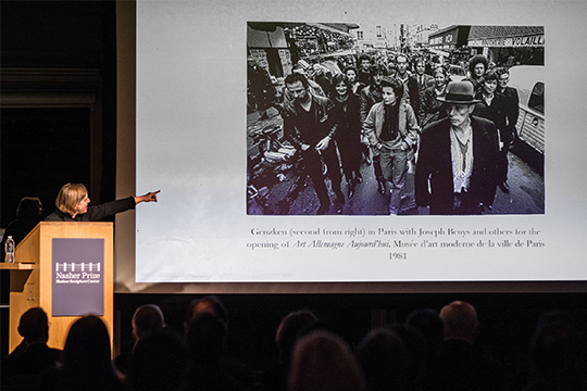 The Keynote Speaker gestures to a screen showing an image of 2019 Nasher Prize Laureate Isa Genzken walking down a street.
