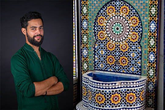 Artist Youssef El Berrichi stands with arms crossed next to an intricate mosaic