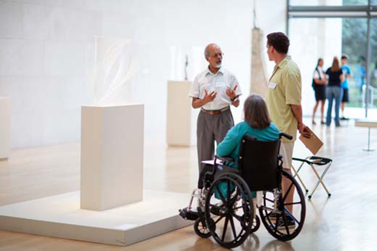 A group of visitors, including one in a wheelchair, discuss a sculpture.