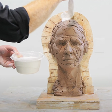 A man applies liquid plaster to a clay mold of a a woman's head