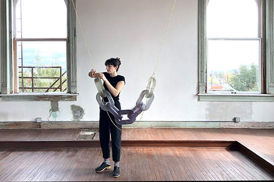 woman in black outfit adjusts a hanging sculpture of links in an artist studio