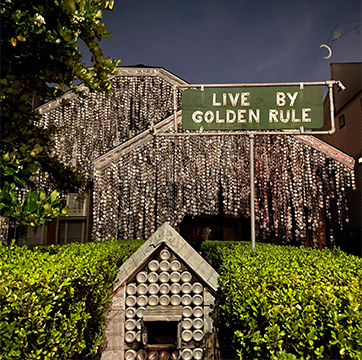 house covered in beer cans with a road sign posted in the yard that says live by golden rule